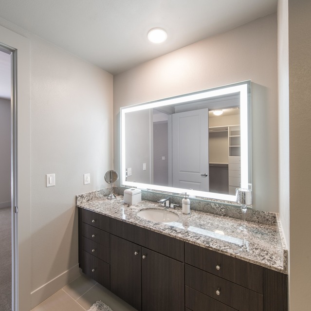 The Arcadian - Bathroom with a Dark Wood Vanity, Granite Countertop, Illuminated Mirror, and a Soft Rug on Tiled Floor