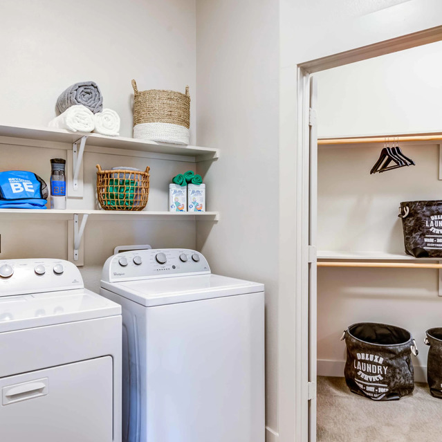The Arcadian - Laundry Room With a White Washer and Dryer, Shelves Holding Folded Towels and Baskets